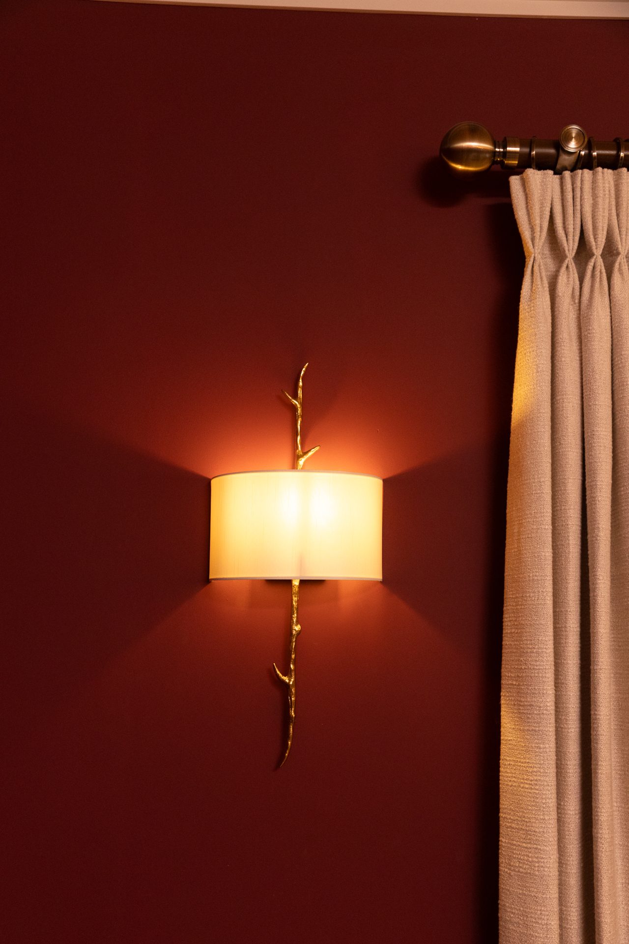 A lit wall sconce with a cream drum shade and a gilded branch-motif backplate, glowing warmly against a deep russet wall, a pleated taupe curtain to the right