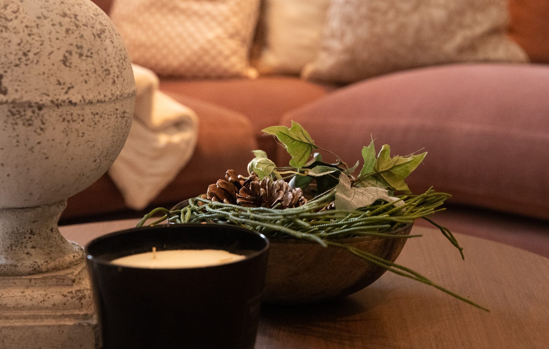 A weathered stone urn finial, a small black candle and a wooden bowl of pinecones and ivy on a round wooden coffee table, terracotta sofa behind, header image for the Late Light chapter