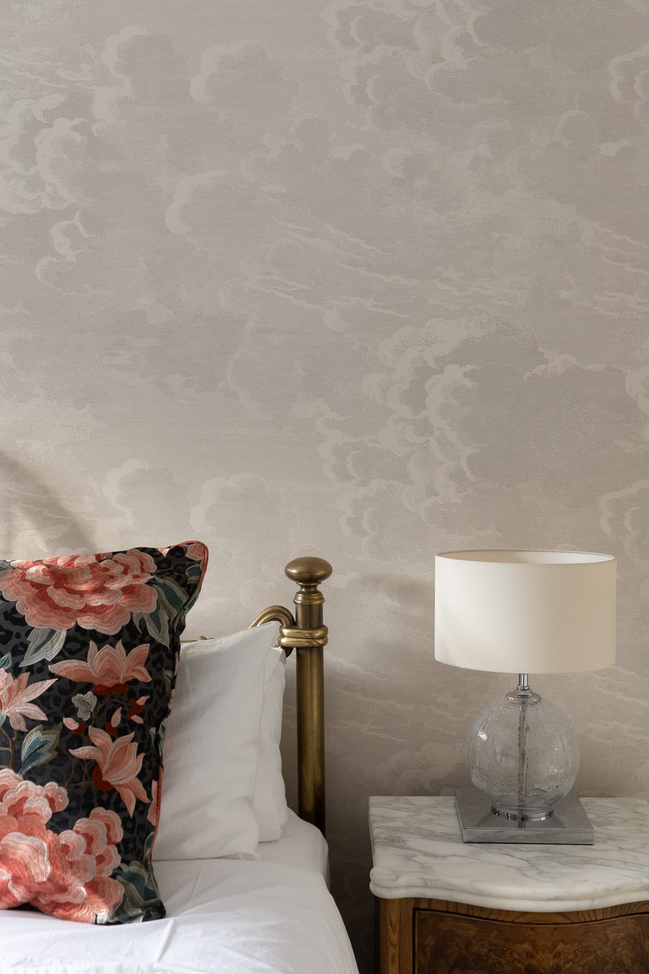 A bedside vignette against soft cloud-pattern wallpaper, a dark floral peony cushion leaning against a brass bed, a bubble-glass lamp with a white drum shade on a marble-topped walnut bedside table
