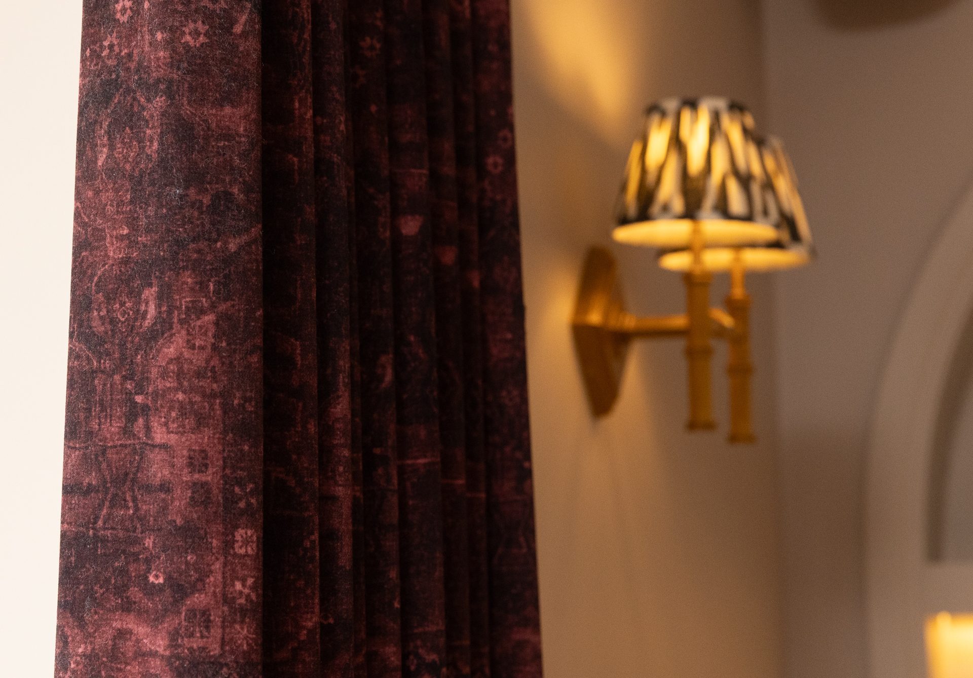 A plum printed curtain with a soft aged-tapestry motif, beside an ornate twin-arm brass sconce with patterned fabric shades glowing against a cream wall