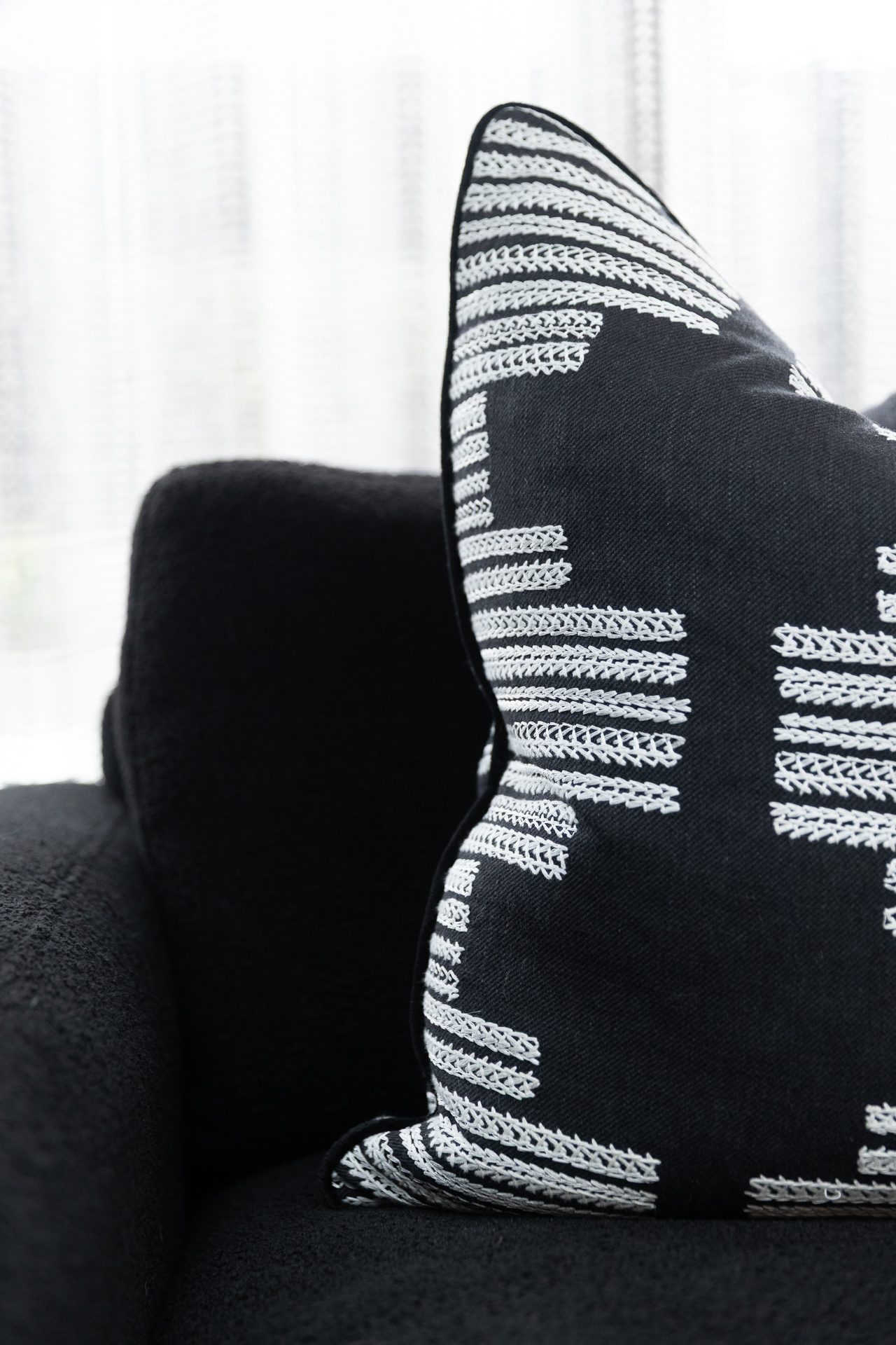 A close-up of a black cushion with white embroidered pattern, resting on a black bouclé armchair against sheer white curtains