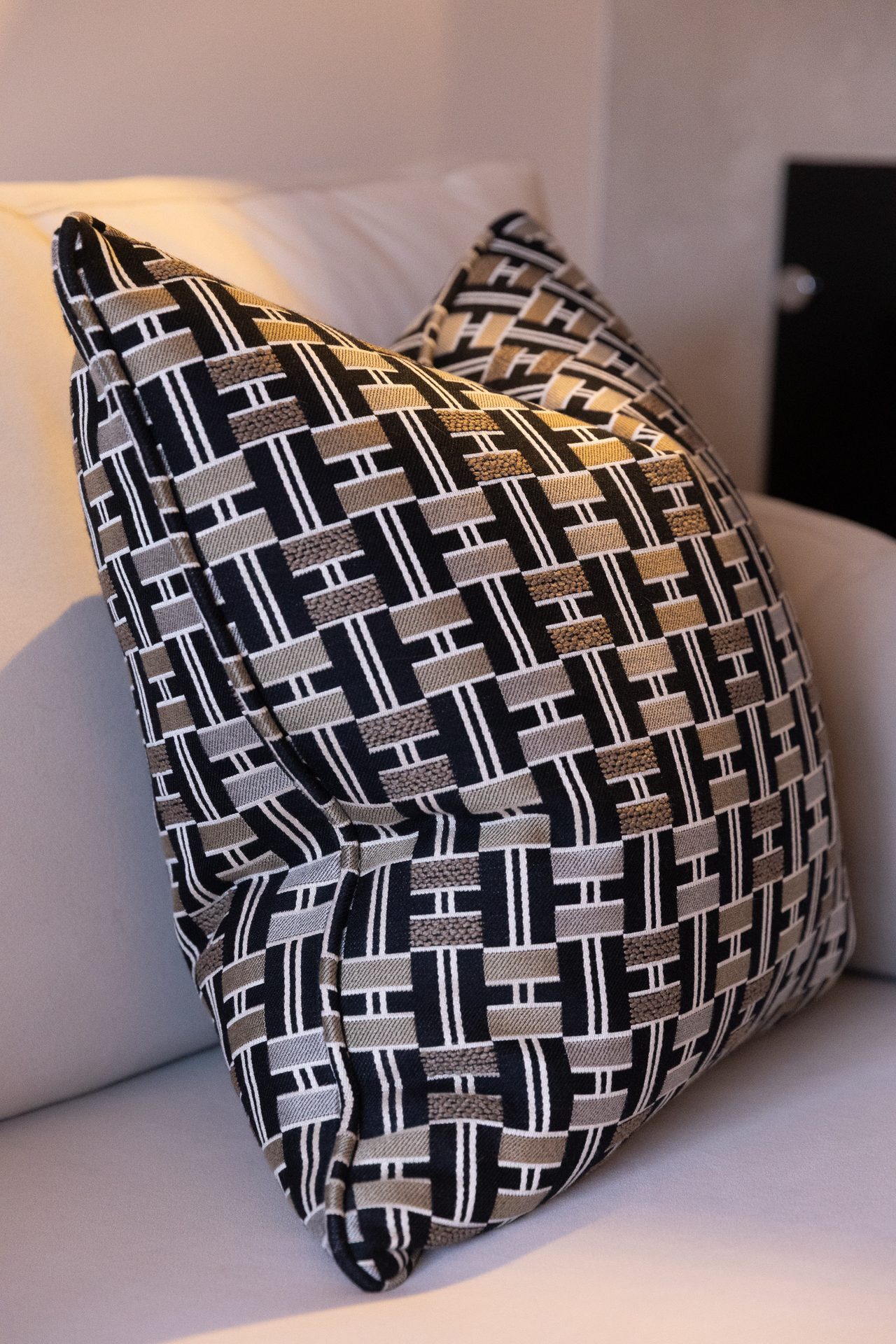 A graphic geometric cushion in black, tan and cream, with an interlocking basket-weave motif, resting on a pale sofa