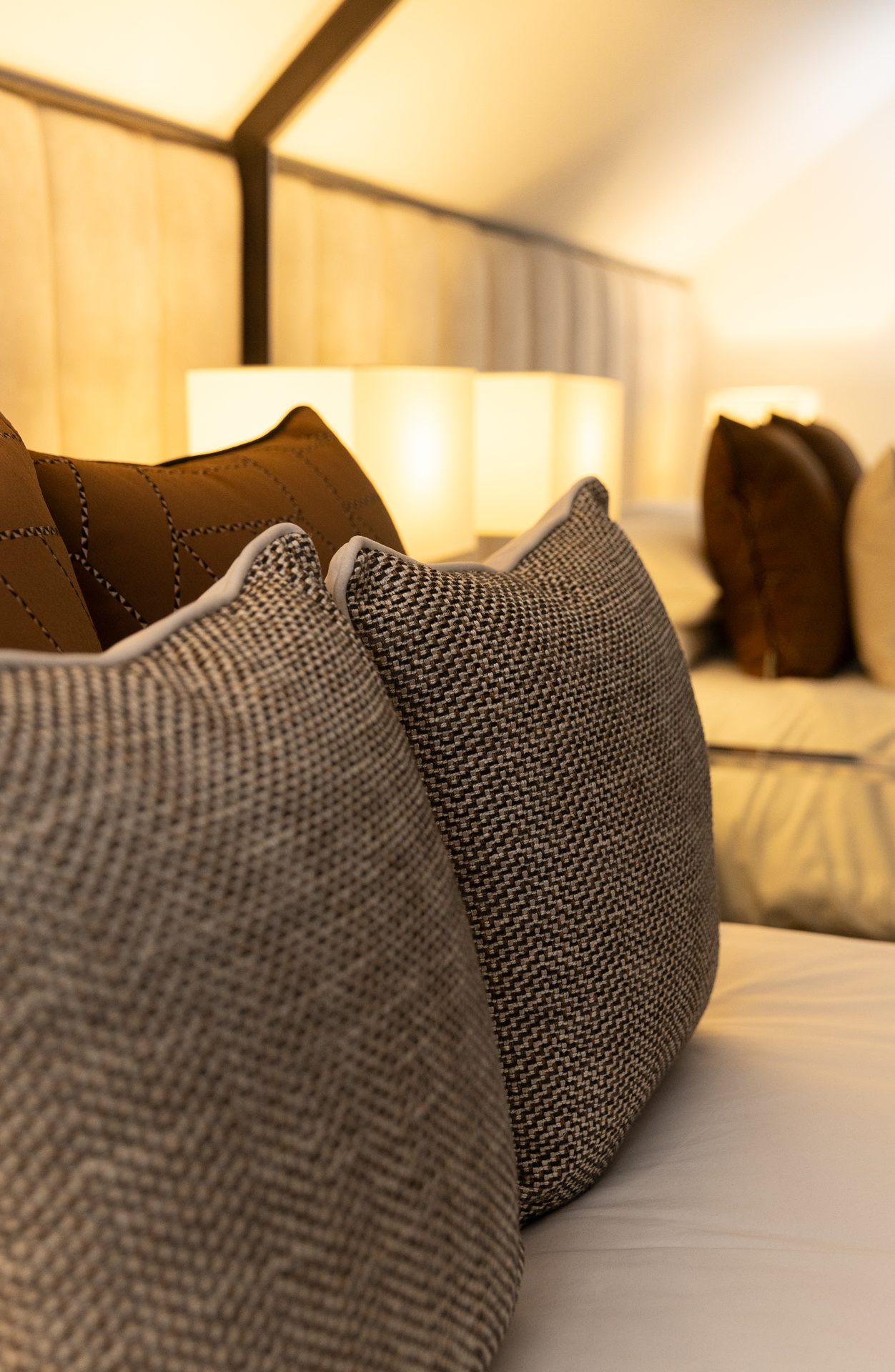 A pair of greige tweed cushions on a white bed, with a tan leather cushion and twin lamps glowing in the background