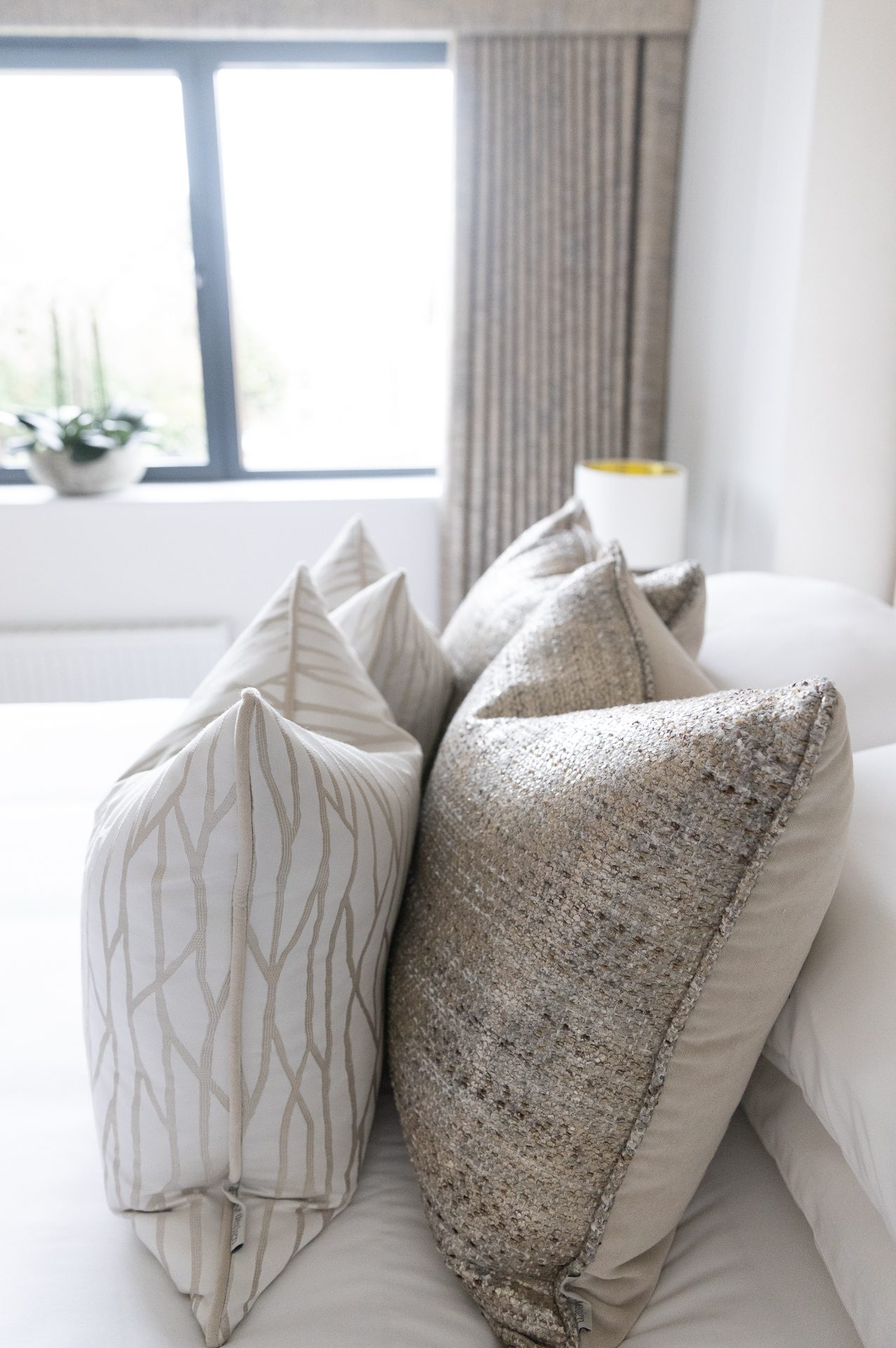 A bedroom corner with cream linear-pattern cushions and a silvered bouclé cushion layered on white bedlinen, a pleated curtain and window behind, header image for the Woven chapter