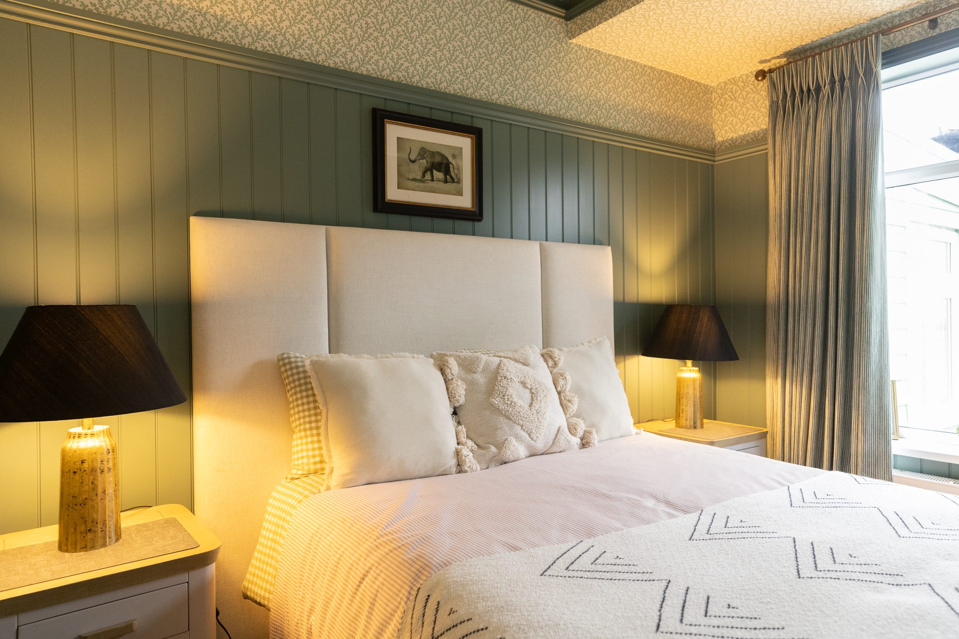 The Bamburgh master bedroom, sage panelling, a deep upholstered bed in white cotton with a tasselled throw and a framed print above