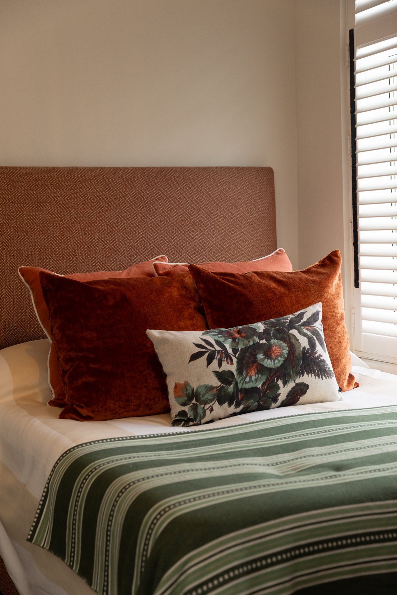 A Bamburgh guest bedroom, a patterned upholstered headboard in mushroom and cream, rust velvet cushions and a botanical lumbar pillow, a green stripe throw, white plantation shutters at the window