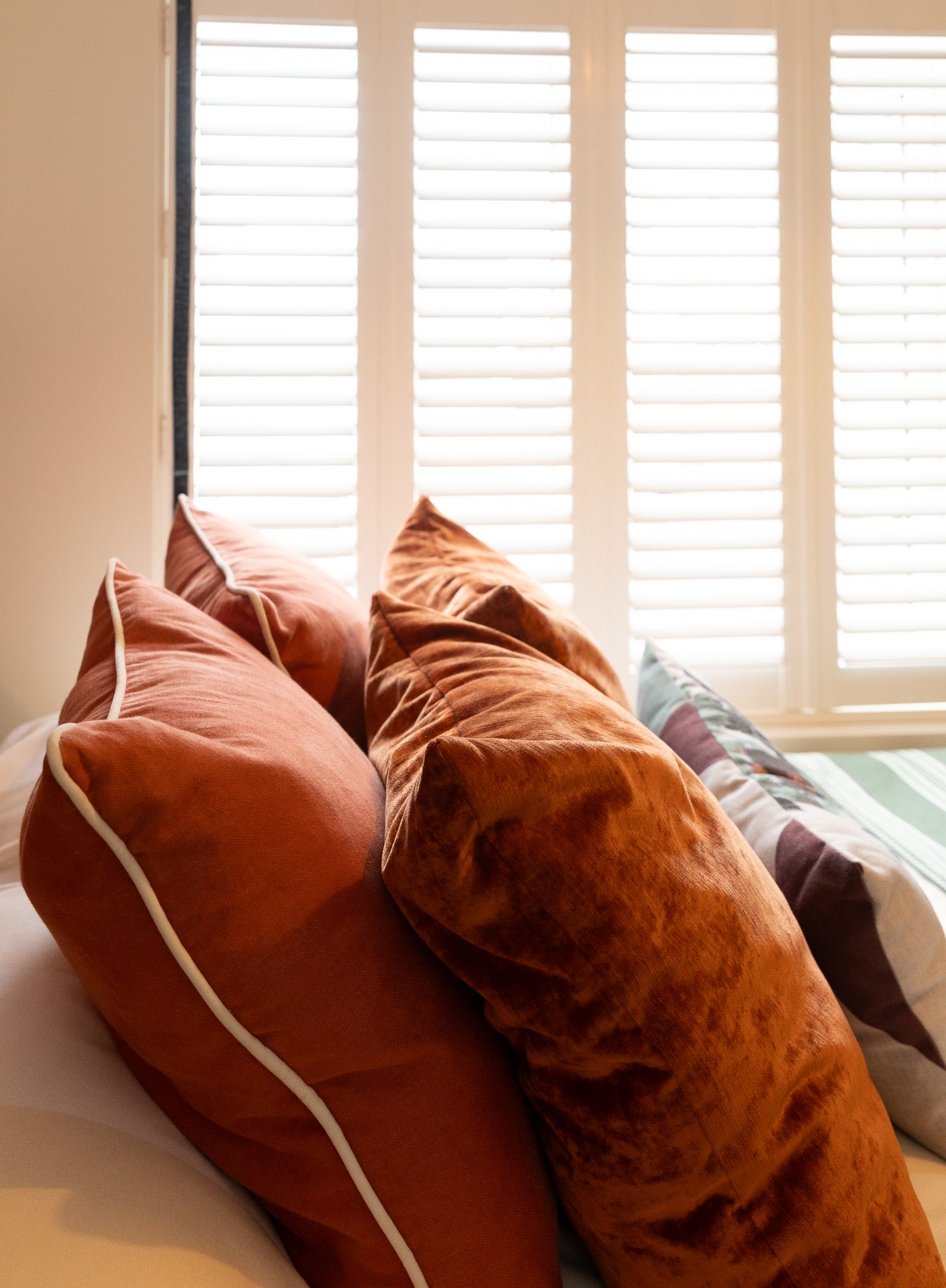 Rust velvet cushions piled against the patterned headboard, white plantation shutters letting in soft light
