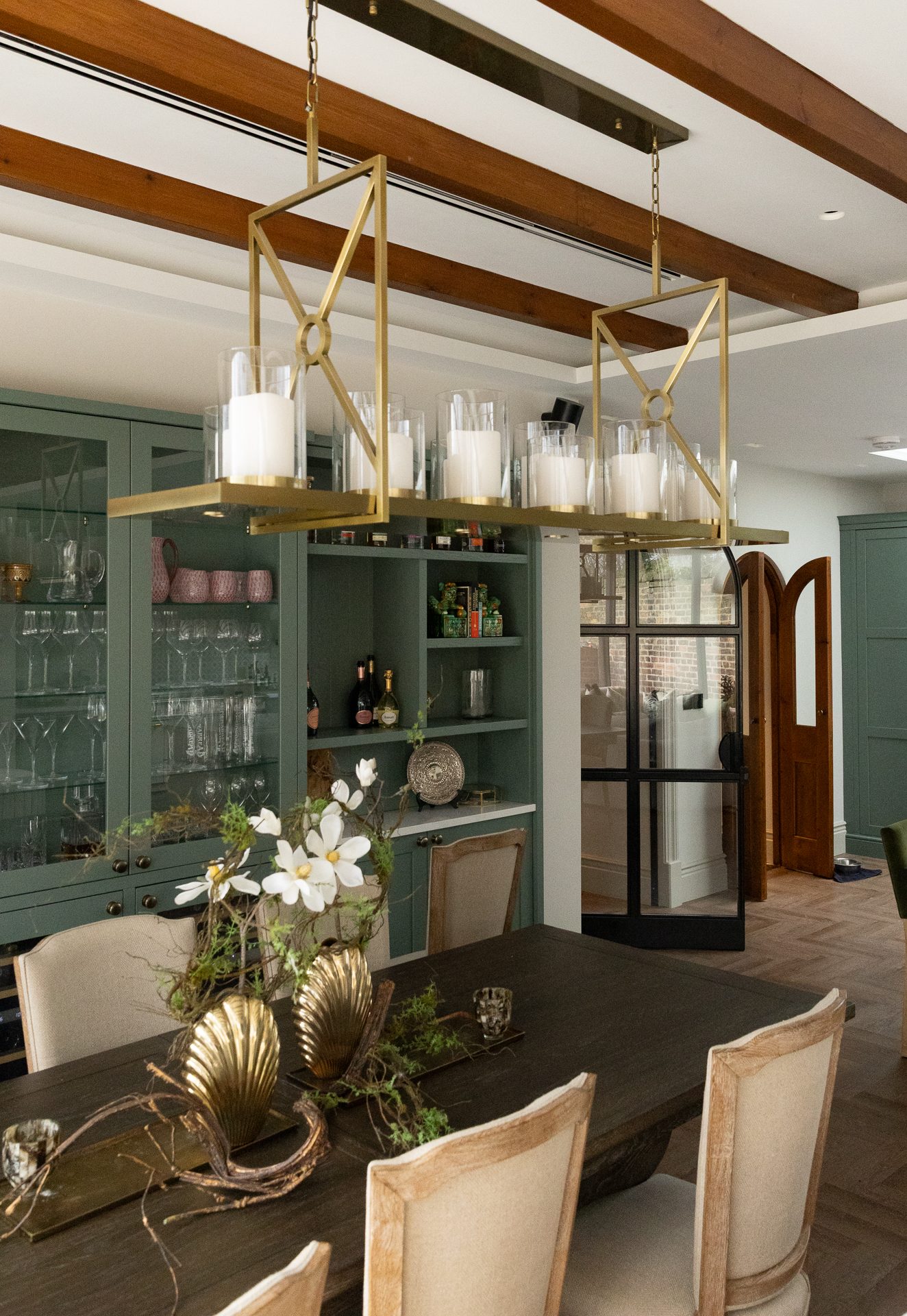 The dining area with brass geometric pendant light, timber beams, and sage green display cabinetry