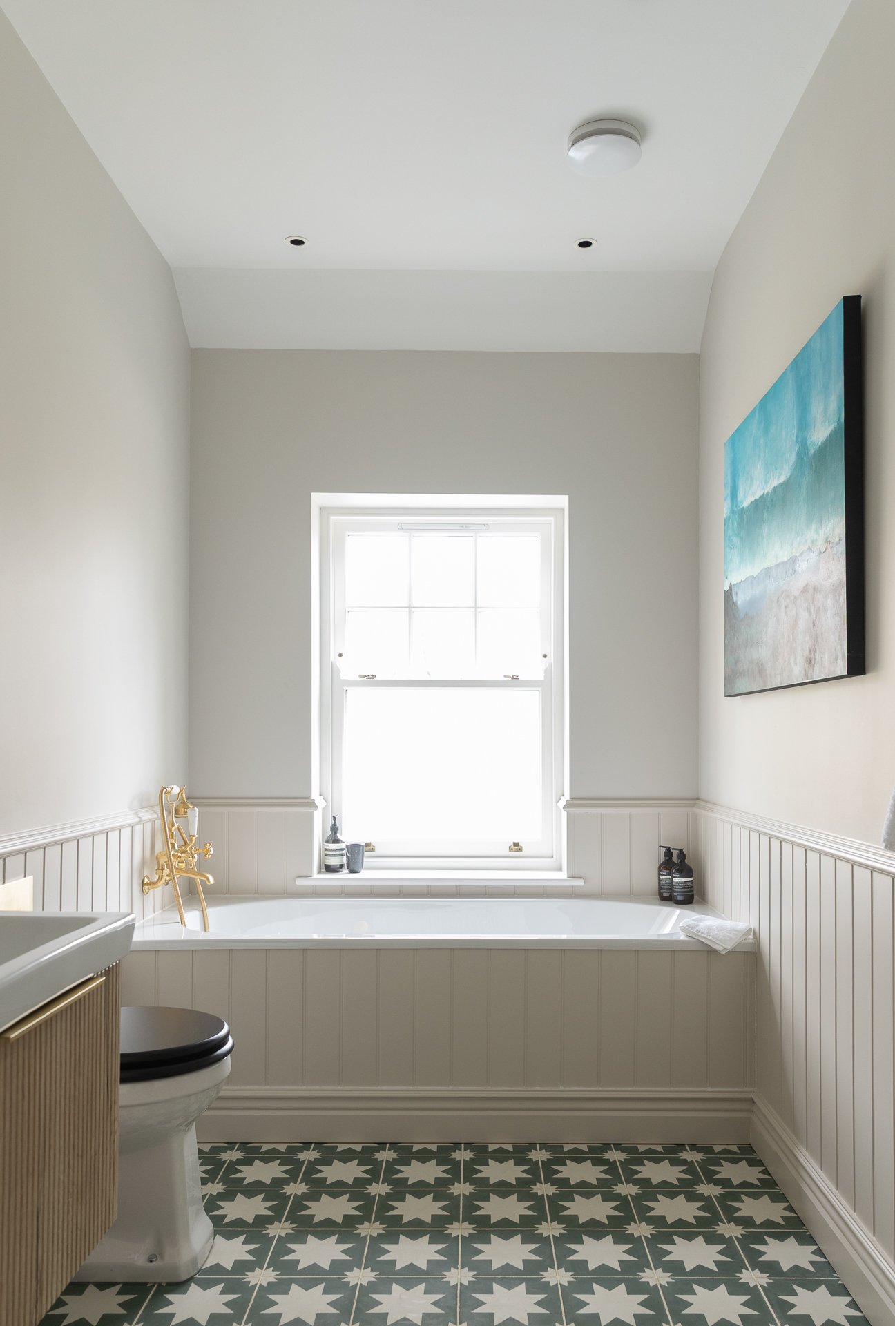 The Elmfield Road family bathroom, a built-in bath below a sash window with cream tongue-and-groove wainscoting, brass wall-mounted tap, reeded timber vanity to the side, framed ocean artwork, and a six-pointed star encaustic tile floor in green and cream
