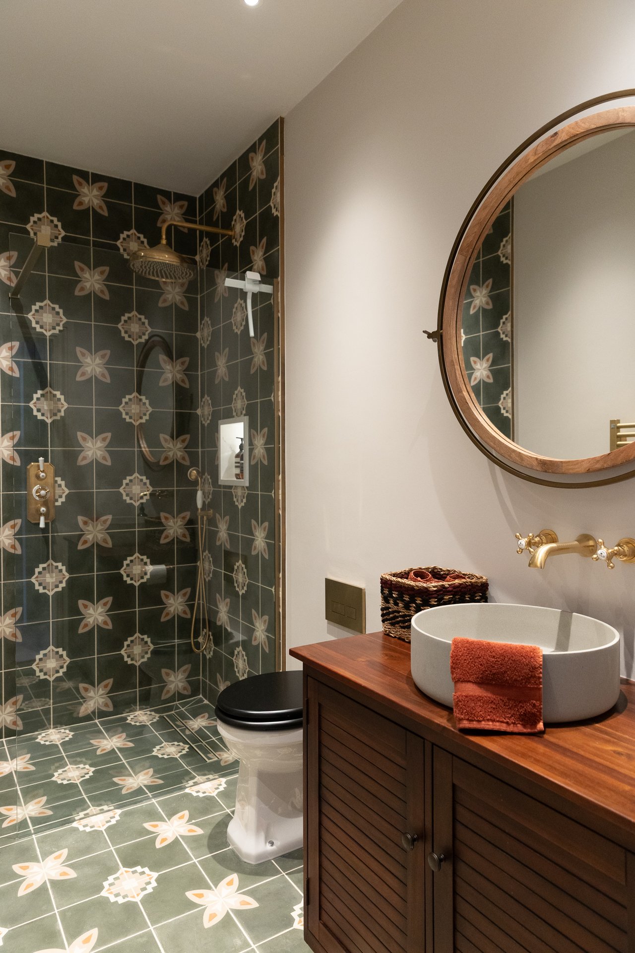 The Elmfield Road shower room, a walk-in shower tiled in deep green encaustic with cream and terracotta floral pattern, brass rain shower and hand shower, a round walnut mirror and slatted vanity with a stone basin and brass wall-mounted tap to the side