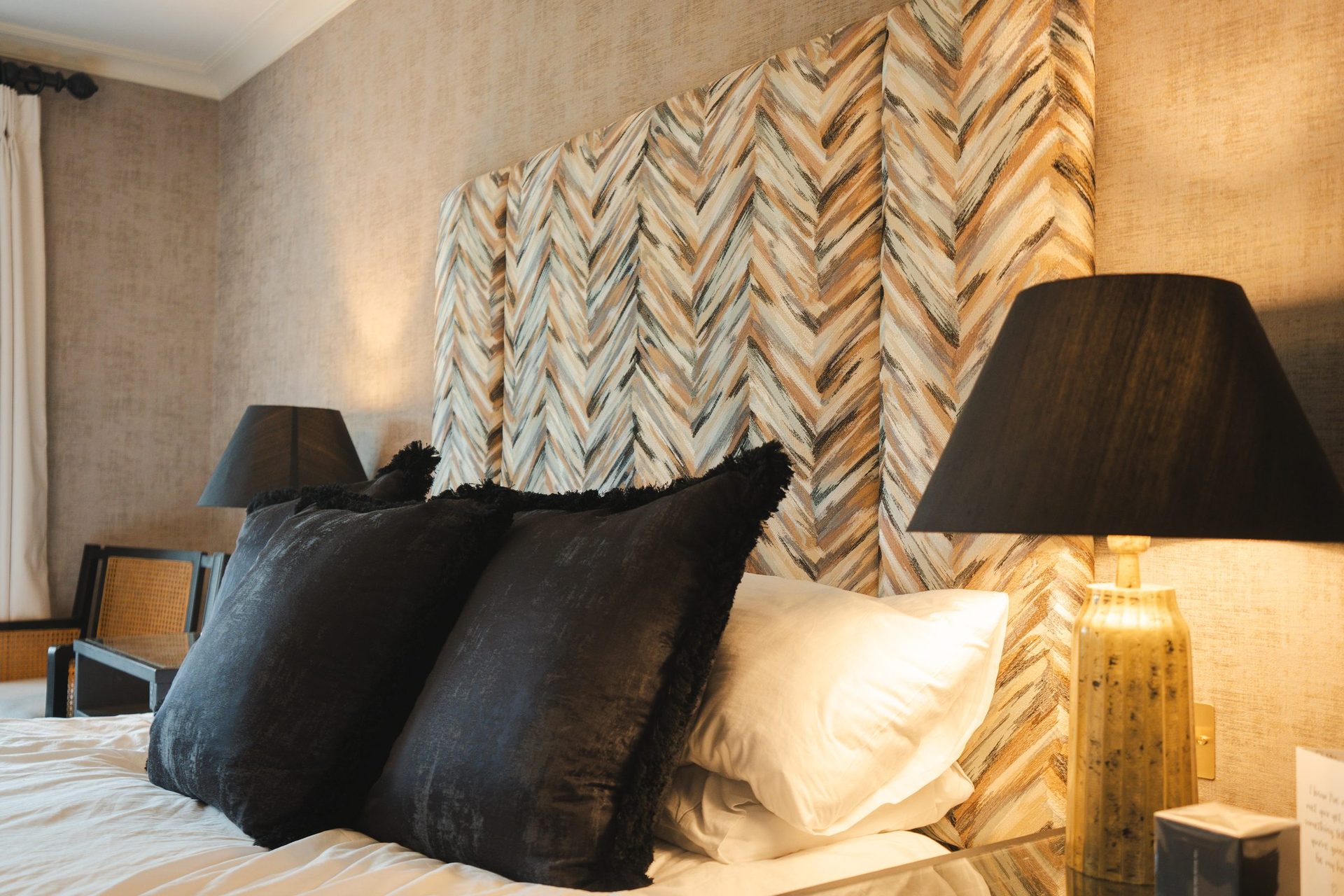 The Montagu bedroom bed, textured chevron headboard, black and cream pillows, a gold lamp with a warm shade on the bedside