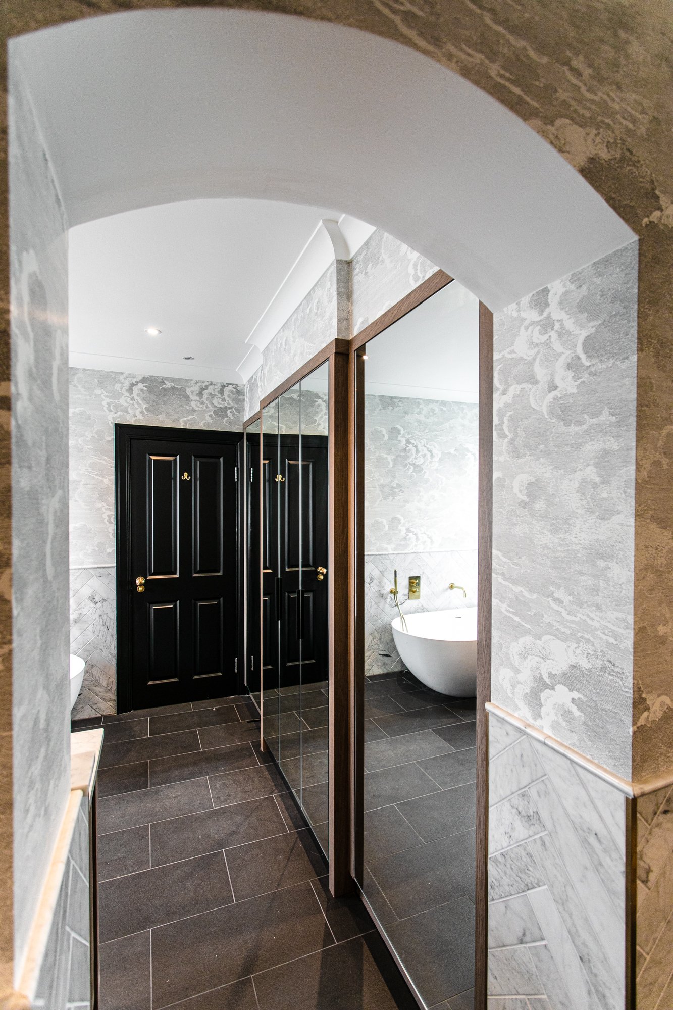 An arched doorway into the master bathroom, Cole and Son cloud wallpaper either side, mirrored wardrobes reflecting a glimpse of the oval bath and marble surround beyond