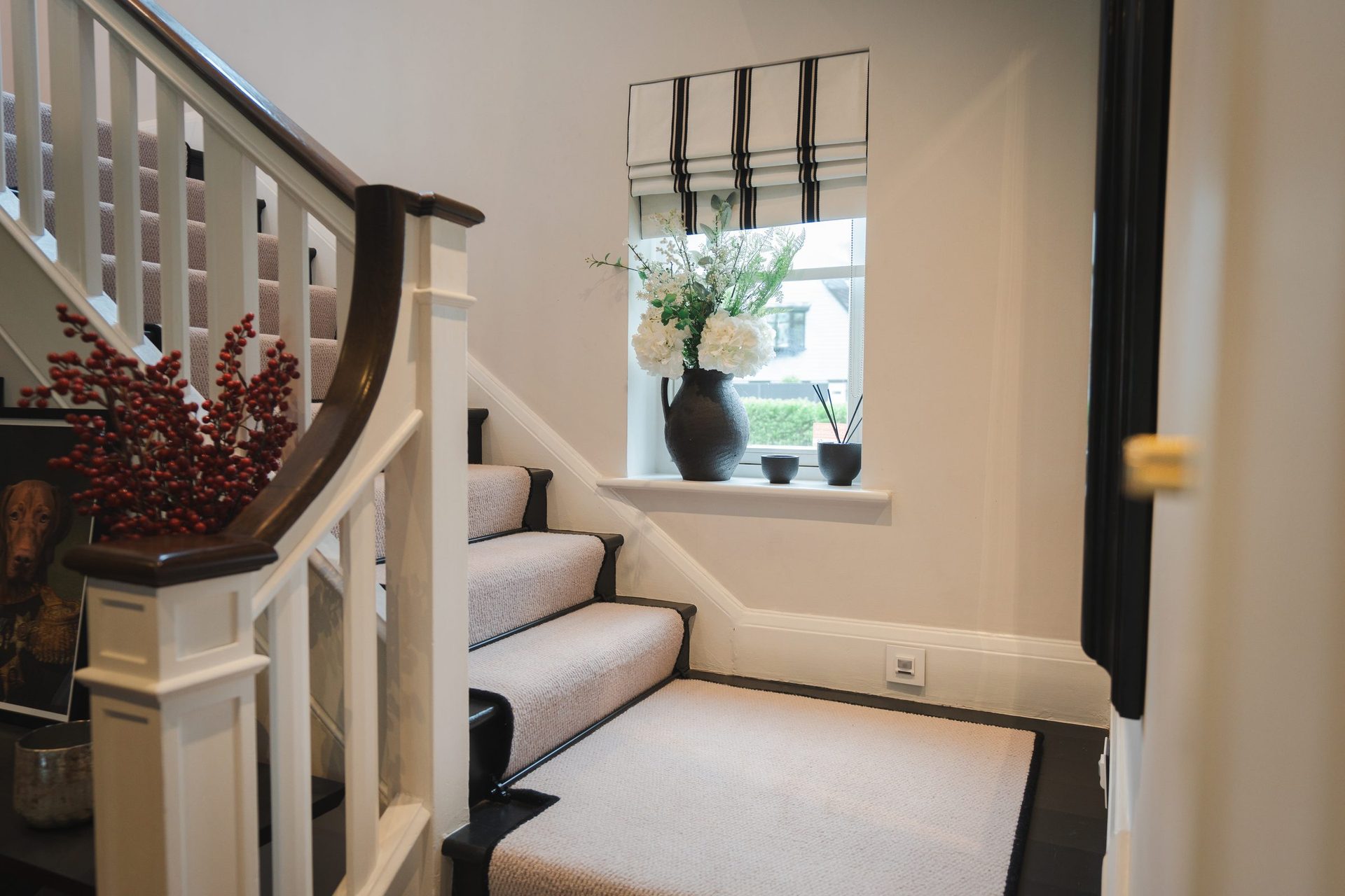 The Montagu staircase, cream runner with black nosing, walnut bannisters, striped Roman blind in the window, console with vase of red berries at the foot