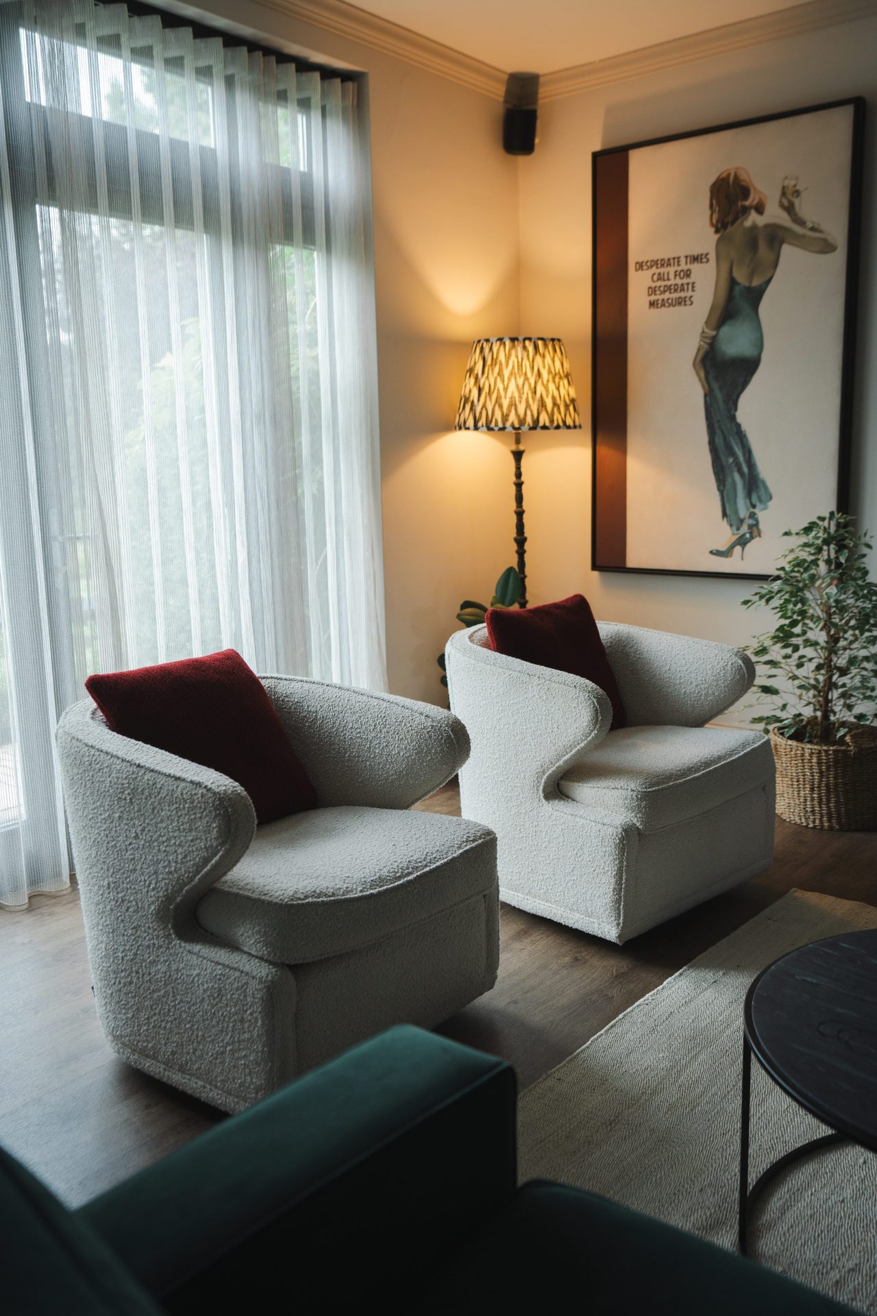 The sitting room held wider, full-length curtain, pair of bouclé armchairs, a potted palm in the corner