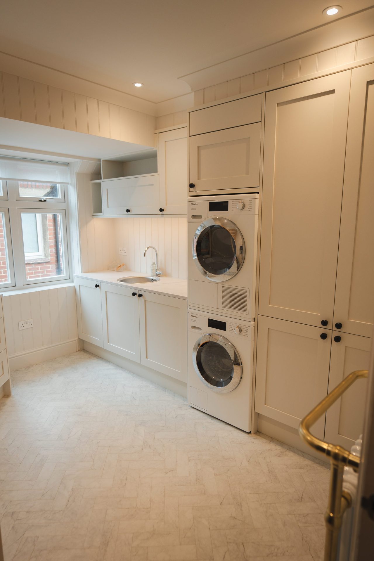The Montagu utility room, cream shaker panelling, stacked washer and dryer, stone herringbone floor, brass hardware