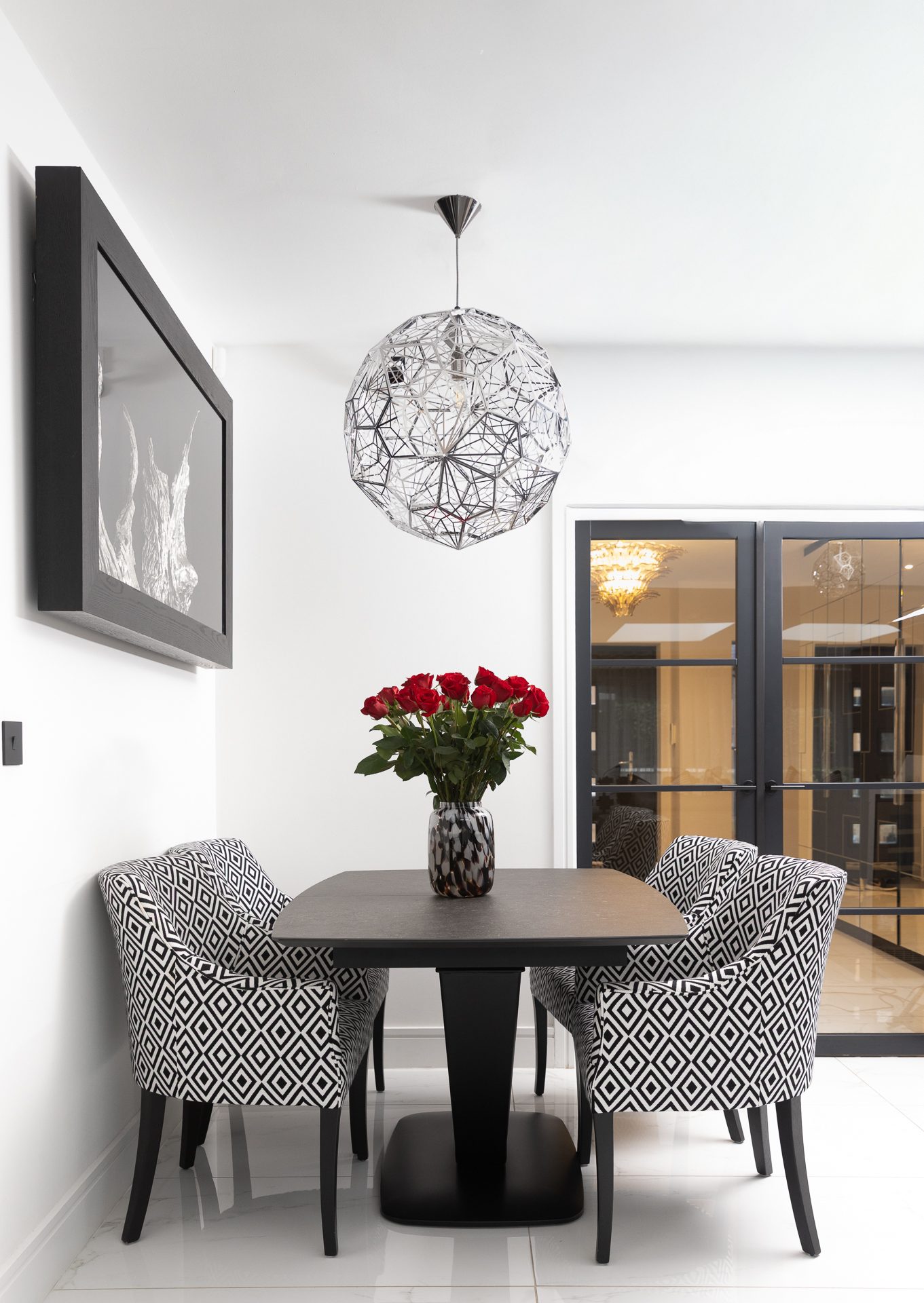 The Sycamore dining room, chrome geometric pendant over a black table, red roses and diamond-pattern chairs with Crittall doors leading through to the hall