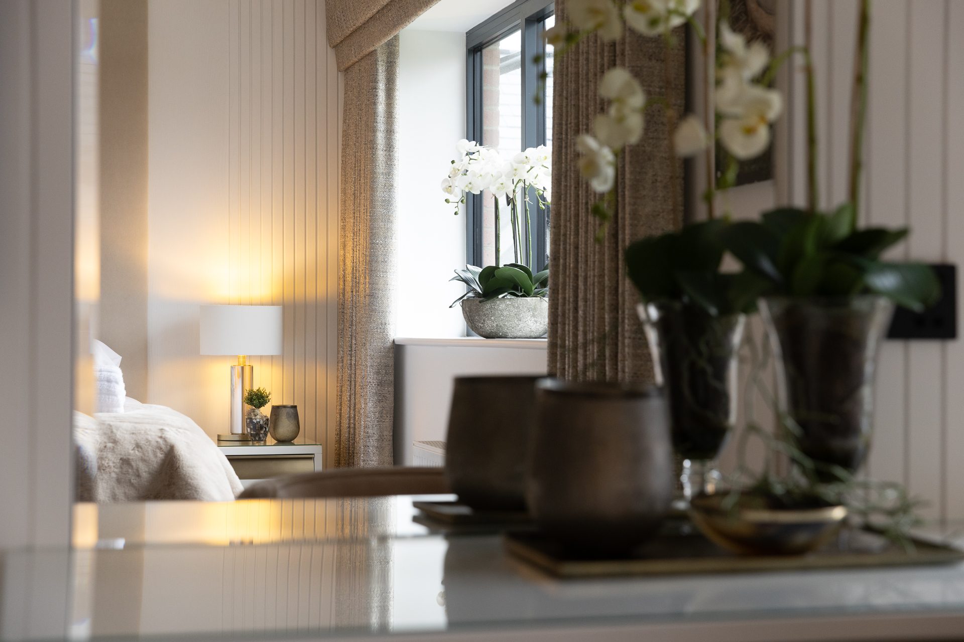 A tableau through the mirror, bedside lamp lit and pewter planters against linen curtains