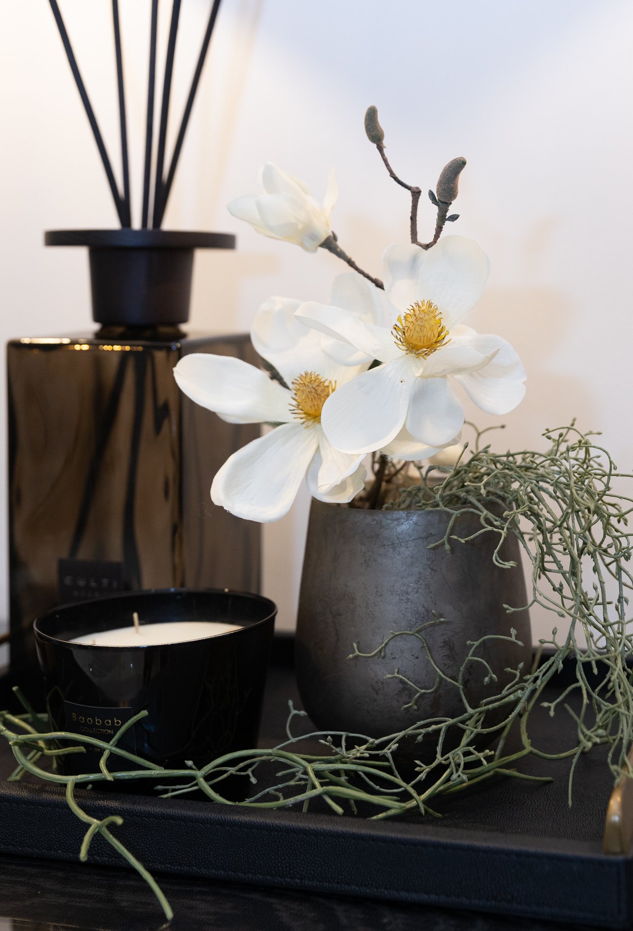 A single white magnolia close up, sitting in a pewter vase with trailing eucalyptus against a dark oak shelf