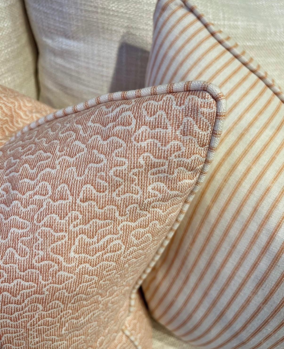 A pair of cushions — one with an ornate pink damask pattern, the other a striped pink and cream ticking — close detail on a cream sofa, in the Andrew Martin signature