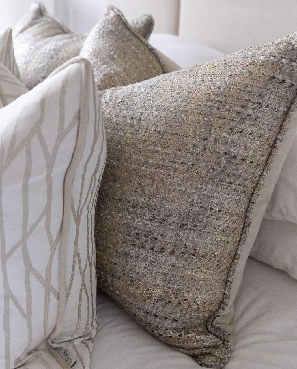 A stack of textured boucle and tweed cushions in cream and grey on a softly upholstered bed — the layered upholstery JAB Anstoetz is known for