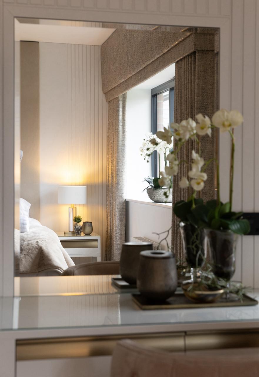 A large floor-standing mirror reflecting a softly lit bedroom, with bronze ceramic vases and white orchids on a glass console — Libra accent and atmosphere pieces in a finished scheme